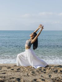 Side view of ethnic female leaning back while standing in ashta chandrasana pose during yoga practice on sandy coast against ocean