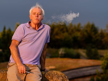 Portrait of man holding electronic cigarette while standing against trees and sky