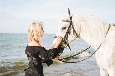 Young blonde woman in black clothes riding white horse on seascape background