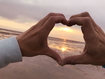Cropped image of hands making heart shape against sea during sunset