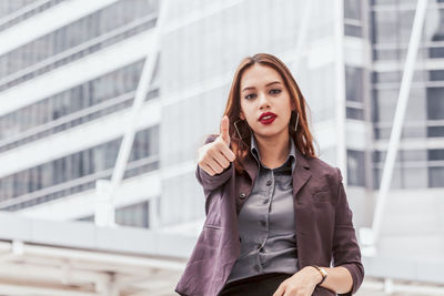 Portrait of young businesswoman showing thumbs up against buildings in city