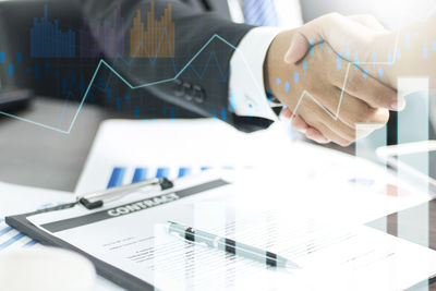 Digital composite image of colleagues giving handshake and line graph at desk in office