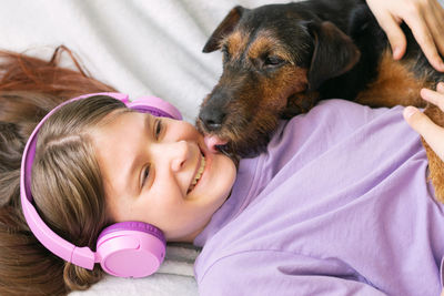 Smiling girl listening music through wireless headphones lying by dog on bed at home