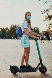 Full length of girl wearing mask standing on electric push scooter on street