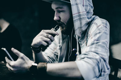 Young man smoking electronic cigarette while using phone at table