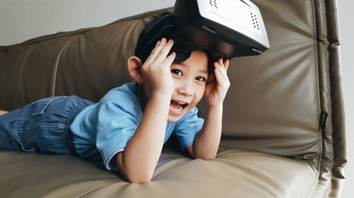 Cheerful boy with virtual reality simulator lying on leather sofa at home