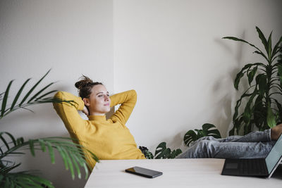 Smiling woman with hands behind head relaxing while sitting by table at home