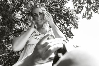 Low angle portrait of young woman holding plant against trees