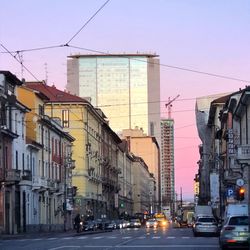 Traffic on city street by buildings against sky during sunset