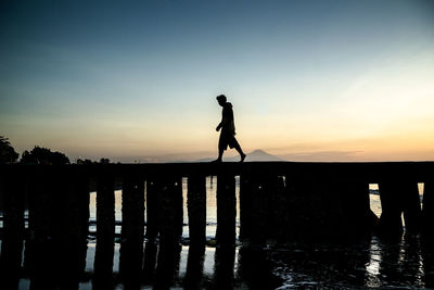 Silhouette man standing by wooden post against sky during sunset