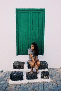 Woman sitting on steps against closed window amidst wall