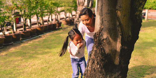 Girl standing with mother behind tree trunk at park