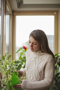 Young woman watering plants while standing against window