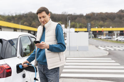 Man using smart phone standing by electric car at station