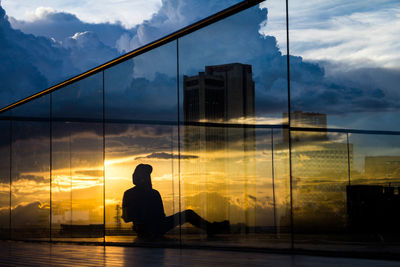 Silhouette man sitting by glass window against sky during sunset