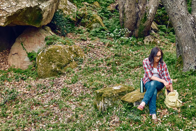 Woman sitting in forest on stone to rest, traveling with backpack in a grove or exercising outdoors.