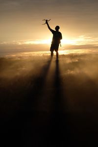 Silhouette man holding drone standing against sky during sunset