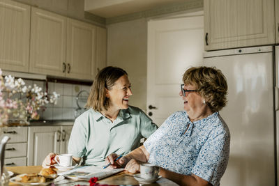 Happy female nurse and senior woman spending leisure time solving crossword puzzle at home