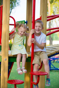 Full length of smiling sibling sitting at play equipment at park