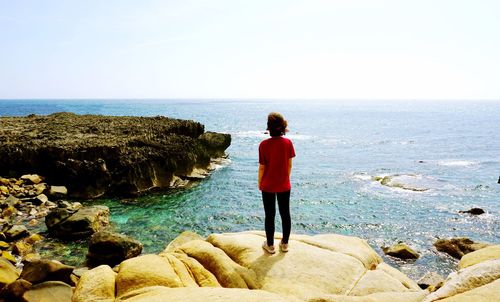 Rear view of woman standing on cliff by sea against clear sky