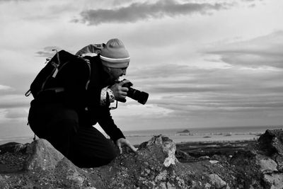 Male hiker photographing at beach against cloudy sky