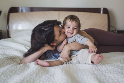 Pretty mom hugging and kissing happy year old baby on white bedspread