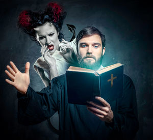 Digital composite image of scared female demon hiding behind priest reading bible