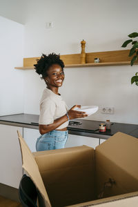 Side view portrait of smiling woman holding bowl while standing near cardboard box in kitchen at home