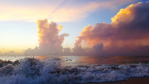 Waves splashing on shore against cloudy sky during sunset