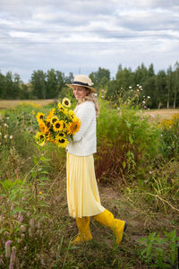 A cute girl in a brimmed hat and yellow rubber boots holds a bouquet of sunflowers in a field