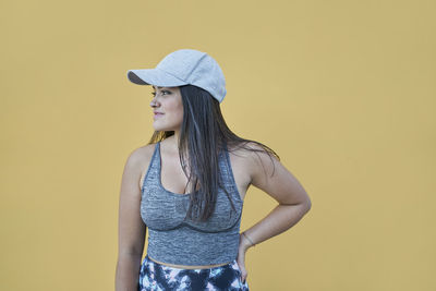 Happy young female in sports top and cap standing with hand on waist against dark yellow wall and looking away with radiant smile