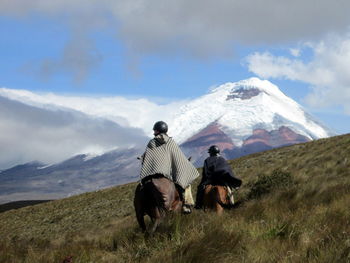 Rear view of people riding horses on grassy field against cotopaxi