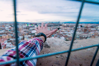 Midsection of person relaxing by window in city against sky