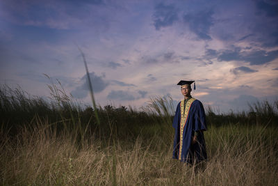 Man in graduation gown standing on field against sky