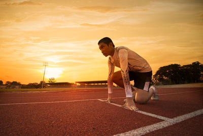 Full length of teenage boy kneeling on sports track against orange sky