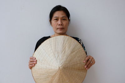 Portrait of mature woman holding asian style conical hat while standing against white wall