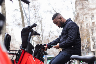 Side view of young masculine black male watching cellphone while using bike parking application in town