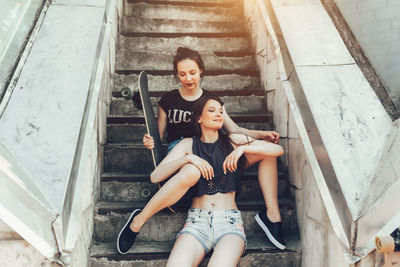 Smiling woman sitting with friend holding skateboard on steps