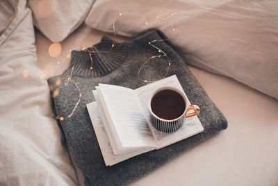 Cup of coffee on knitted textile sweater in bed with paper open book with christmas light closeup