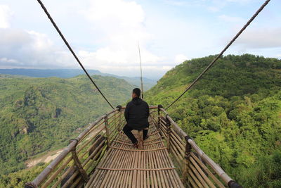 Rear view of man looking at lush mountains while sitting on observation point