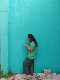 Young woman using mobile phone while standing against blue wall