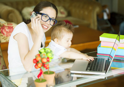 Mother holding cute baby son while talking on mobile phone at home