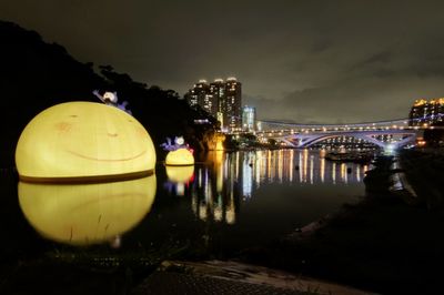 Illuminated bridge over river by buildings against sky at night