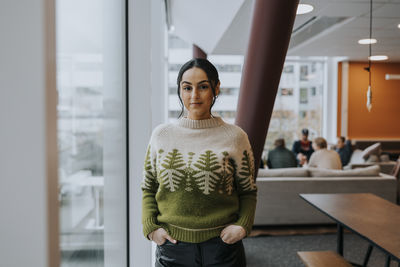 Portrait of confident businesswoman with hands in pockets standing by window at office