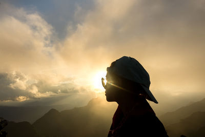 Woman in cap and sunglasses standing against sky during sunset