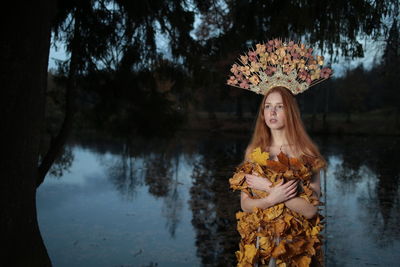 Thoughtful young woman covered with leaves standing by lake at forest during autumn