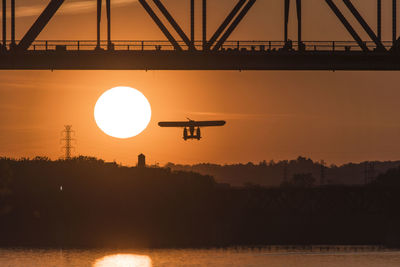 Silhouette airplane flying by george rogers clark memorial bridge over ohio river against sky during sunset