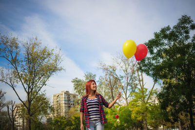 Low angle view of smiling woman with balloons standing against sky in park