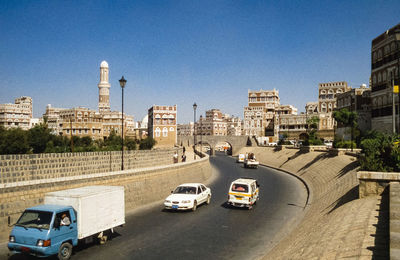 Cars on road by buildings against clear blue sky