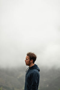 Young man looking away while standing on snow against sky